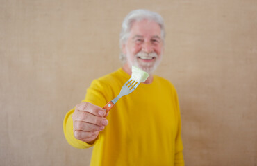 Wall Mural - Defocused white-haired senior man in yellow shirt isolated on a light background holds a fork with a piece of melon ready to be eaten