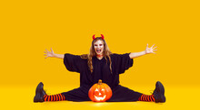 Funny Crazy Screaming Woman With Fake Neck Blood Cut In Black Halloween Costume, Red Devil Horns And Striped Stockings Does Leg Split On Floor With Jack O Lantern Pumpkin On Yellow Studio Background