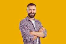 Studio Shot Of A Happy Young Guy. Cheerful Handsome Bearded Young Man Wearing A Casual Shirt Standing With His Arms Folded Isolated On A Yellow Color Background, Looking At The Camera And Smiling