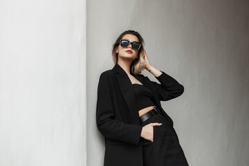 Elegant fashionable business glamour woman model with fashion sunglasses in black clothes with top and stylish black blazer stands near a wall on the street