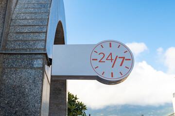 A white banner with the image of a clock signifying the round-the-clock operation of the institution against the background of a blue sky. twenty-four by seven round-the-clock work signage