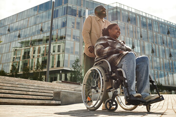 Wall Mural - Low angle portrait of smiling black man assisting partner in wheelchair and enjoying walk together in city against glass architecture, copy space