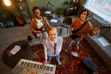 Wall Mural - High angle portrait of music band practicing in home studio with focus on young blonde woman holding microphone and looking up at camera