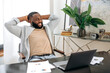 © Kateryna - Happy calm african american man, freelancer, company employee, sits at the workplace in the office, takes a break from work, puts his hands behind his head, looks to the side, dreams of rest, smiles