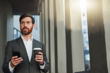 Sticker - Focused european businessman with coffee and phone near office building during break looking away