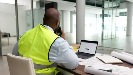 Canvas Print - Young construction manager working on a laptop at his desk while speaking on a communication radio.Professional industry worker planning projects in his office. Contractor putting on his hardhat.