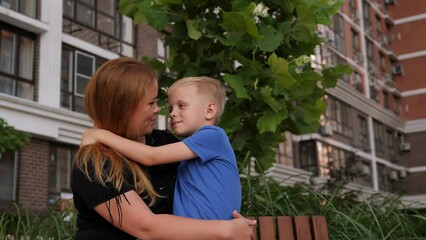 a mother hugs her son sitting on a bench in the yard of a multi-storey building.