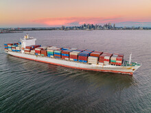 Aerial View Of A Cargo Ship Leaving Port At Sunset, Fully Laden With Containers