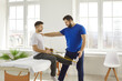 Physiotherapy and rehabilitation after sports injuries. Male physiotherapist assisting man to exercise with resistance band in rehabilitation clinic. Young athlete sitting on couch in hospital office.