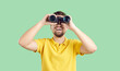canvas print picture - Happy joyful young man in casual yellow T shirt standing isolated on green background, holding modern binoculars, looking ahead and smiling. Making discoveries and having fun while travelling concept