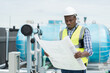 © amorn - African American male engineer working with building blueprint for checks or maintenance sewer pipes or water tank area at construction site on rooftop of building