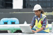 © amorn - Female engineer typing on laptop computer for checks or maintenance in sewer pipes area at construction site. African American woman engineer working in sewer pipes area at rooftop of building