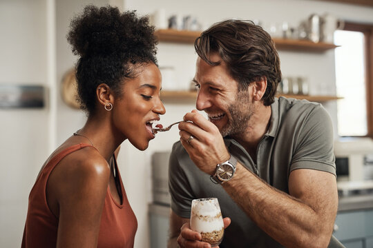 Romantic, happy and interracial couple eating a healthy yogurt together in a cute, sweet and fun kitchen romance. Loving, in love and excited husband feeding his beautiful afro wife delicious dessert