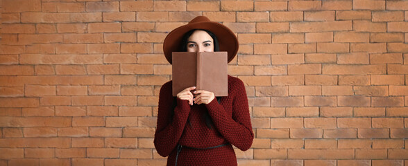 Wall Mural - Stylish young woman in autumn clothes and with book on brick background with space for text