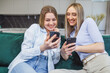 © Ivan - Joyful female friends checking photos on a mobile phone, spending time together at home, Two young ladies are sitting side by side on a sofa in the living room, using a smartphone