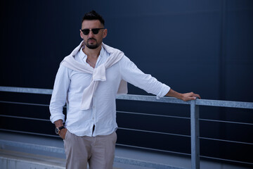 Wall Mural - Cool modern young hipster man with a stylish hairstyle in sunglasses in a white shirt. Attractive urban guy posing near a dark wall. Fashionable spring menswear.