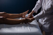 Detalhe das mãos de massagista aplicando massagem terapêutica no pé de um paciente que está deitado em uma maca com lençol branco.