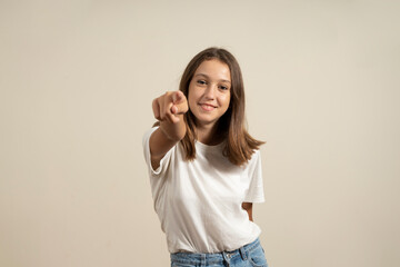 I choose you and command you. The smiling teenage girl pointing at camera, half-length close-up portrait on beige studio background. Human emotions, facial expression concept. Front view.