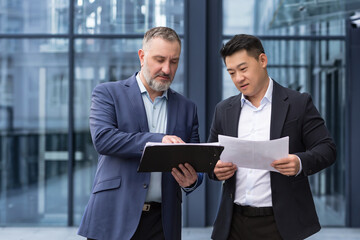 Wall Mural - Team of two businessmen workers outside office building discussing and thinking over documents financial reports and accounts, senior gray haired man and asian colleagues business partners