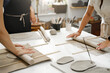 © ninelutsk - Pottery workshop in sunny studio. People working with clay on the table. Adults learning to do ceramic plates. Pottery as hobby and leisure activity. Hands cose-up producing plates