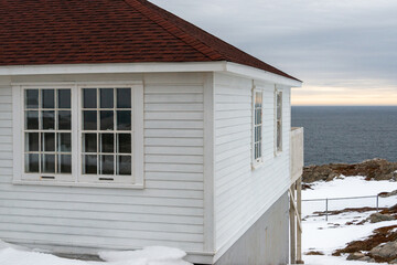 The exterior of a white wooden clapboard building with two glass windows, a black roof, and a wooden patio overlooking a rocky coastline with blue ocean and an orange setting sky in the background. 