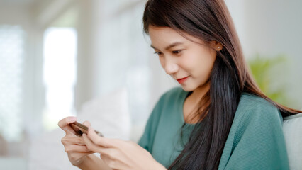 Wall Mural - Happy young asian woman relax on comfortable couch at home texting messaging on smartphone, smiling girl use cellphone chatting, browse wireless internet on gadget, shopping online from home