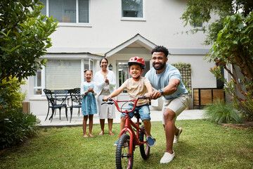 Boy on bicycle learning with proud dad and happy family in their home garden outdoors. Smiling father teaching fun skill, helping and supporting his excited young son to ride, cycle and pedal a bike