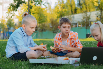 cute caucasian children sitting on grass in park having lunch eating pizza outdoors after school.