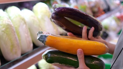 Wall Mural - The customer chooses vegetables to prepare a tasty and healthy dinner for the whole family. A woman takes eggplants packed in a film from a shelf of zucchini for convenience.