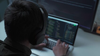 Handsome male software developer programming codes while working behind laptop with headphones from home. IT engineer in headphones working on computer in office. Freelancer works, social distancing.