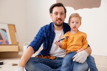 Wall Mural - Cute middle-aged dad sitting on the floor and holding his 2-year-old son on his lap. The child is wearing an orange sweater and jeans, holding a brush in his hand. Around them are repairs.