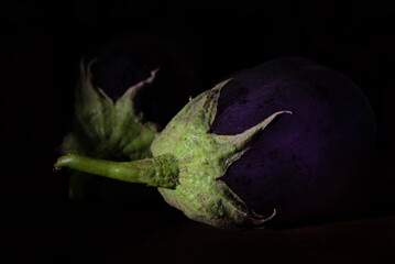 dark shot of two purple aubergines with green stems lying side by side against a dark background.