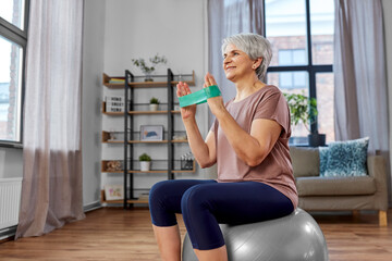 Canvas Print - sport, fitness and healthy lifestyle concept - smiling senior woman with resistance band sitting on exercise ball at home