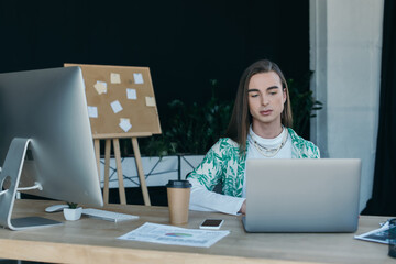 Queer person using laptop near devices and coffee in office.