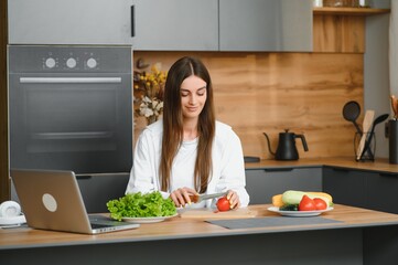 Wall Mural - Athletic woman blogger nutritionist prepare a salad with fresh vegetables and conducts a video conference on healthy eating on laptop in the kitchen