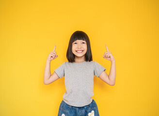 Wall Mural - Portrait of young fun smart happy little cute asian girl isolated on yellow with copy space studio shot. Education for elementary kindergarten, little girl finger point up back to school concept