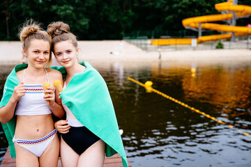 Two teenage girls in swimsuit eating ice cream wrapped in a green towel standing on bridge at summer vacation.