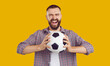 © Studio Romantic - Funny, crazy male football fan supporting his team. Portrait of a happy, excited young man in a casual shirt standing isolated on a yellow color background, holding a soccer ball and screaming loudly