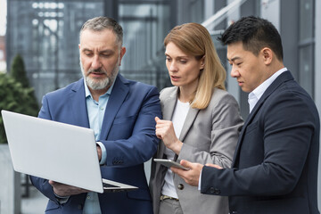 Diverse business group of three people talking and discussing strategy outside office building businessman and businesswoman looking at laptop screen while standing