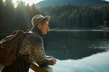 Portrait of young millennial traveller man in hat with backpack enjoying mountain lake with blue water ang forest around