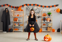 Kid In Spooky Halloween Costume. Portrait Of Child Holding Orange Pumpkin Bucket. Girl Wearing Black Dress, Witch Hat And Scary Makeup Standing In Room Decorated With Jack-o-lanterns And Spider Webs