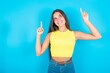 © Jihan - Successful friendly looking brunette woman wearing yellow tank top over blue background exclaiming excitedly, pointing both index fingers up, indicating something.