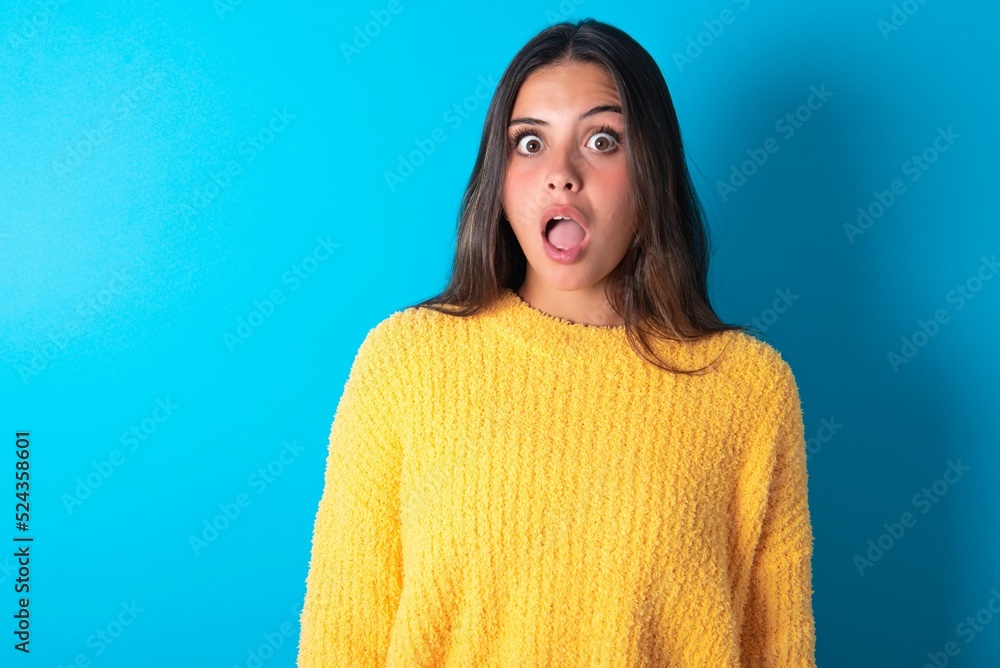 Shocked beautiful brunette woman wearing yellow sweater over blue background stares bugged eyes keeps mouth opened has surprised expression. Omg concept
