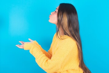 Profile side view view portrait of attractive beautiful brunette woman wearing yellow sweater over blue background sending air kiss