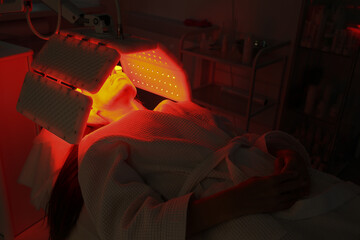 Express facial treatment with led therapy. Beautiful girl on a light therapy procedure. LED lamp with red and yellow light. Safe skin care. Woman in protective glasses. Beauty and wellness concept. 