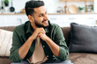 © Kateryna - Handsome positive confident attractive indian or arabian man, in casual stylish clothes, sitting at home in cozy living room on the background of the kitchen, looks to the side, smiling