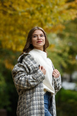 Poster - Close-up portrait of young woman in a stylish trench coat and in a white sweater resting in a park. warm autumn day.