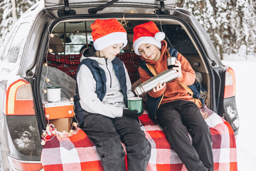 Wall Mural - Two friends teenagers boys in red santa hats with thermos sitting in trunk of car decorated for Christmas. Road trip adventure and local travel concept. Winter picnic in snowy forest New year vacation