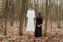 White Wedding Dress Hanging On Line In Woods And Woman In Black Standing Near By