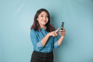 Wall Mural - A portrait of a happy Asian woman is smiling and holding her smartphone wearing a blue shirt isolated by a blue background